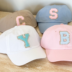 May include: Four baseball caps with different colors and letters on the front. The caps are beige, blue, white, and pink. The letters are C, S, Y, and B. The letters are made of a light blue material with gold trim.