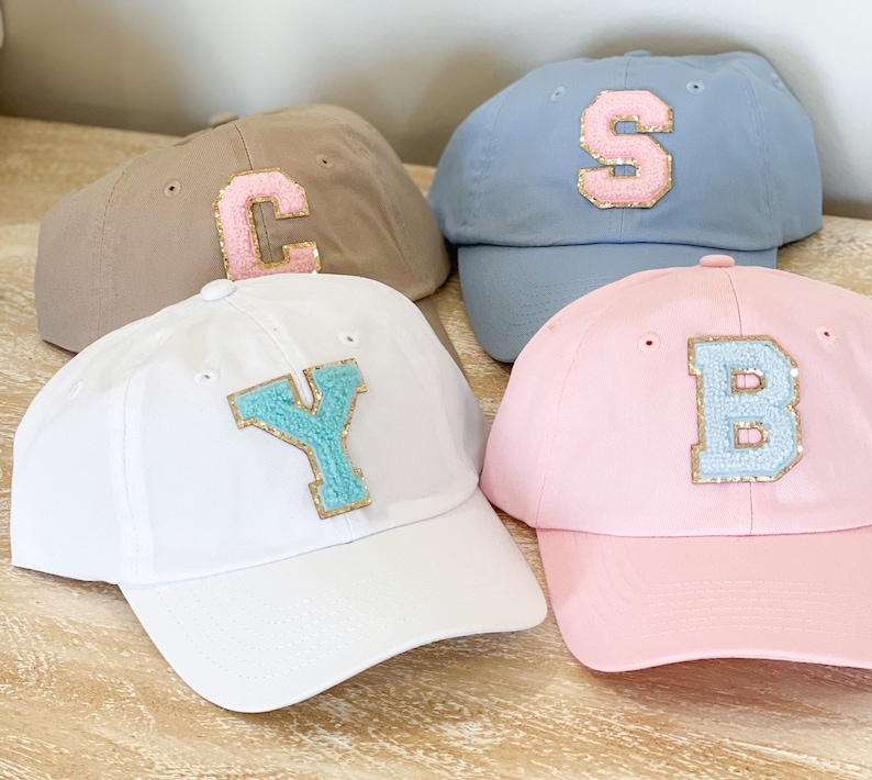 May include: Four baseball caps with different colors and letters on the front. The caps are beige, blue, white, and pink. The letters are C, S, Y, and B. The letters are made of a light blue material with gold trim.