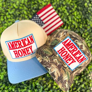 May include: Two baseball caps with the text "AMERICAN HONEY" on a white and blue rectangular patch. One cap is tan with a light blue brim, the other is camouflage. An American flag is in the background.