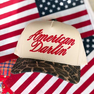 Puede incluir: Una gorra beige tipo trucker con el texto American Darlin' bordado en rojo en el panel frontal. La gorra tiene una corona estructurada y una visera curva con un patr&oacute;n de estampado animal marr&oacute;n y negro, sobre una bandera roja, blanca y azul.