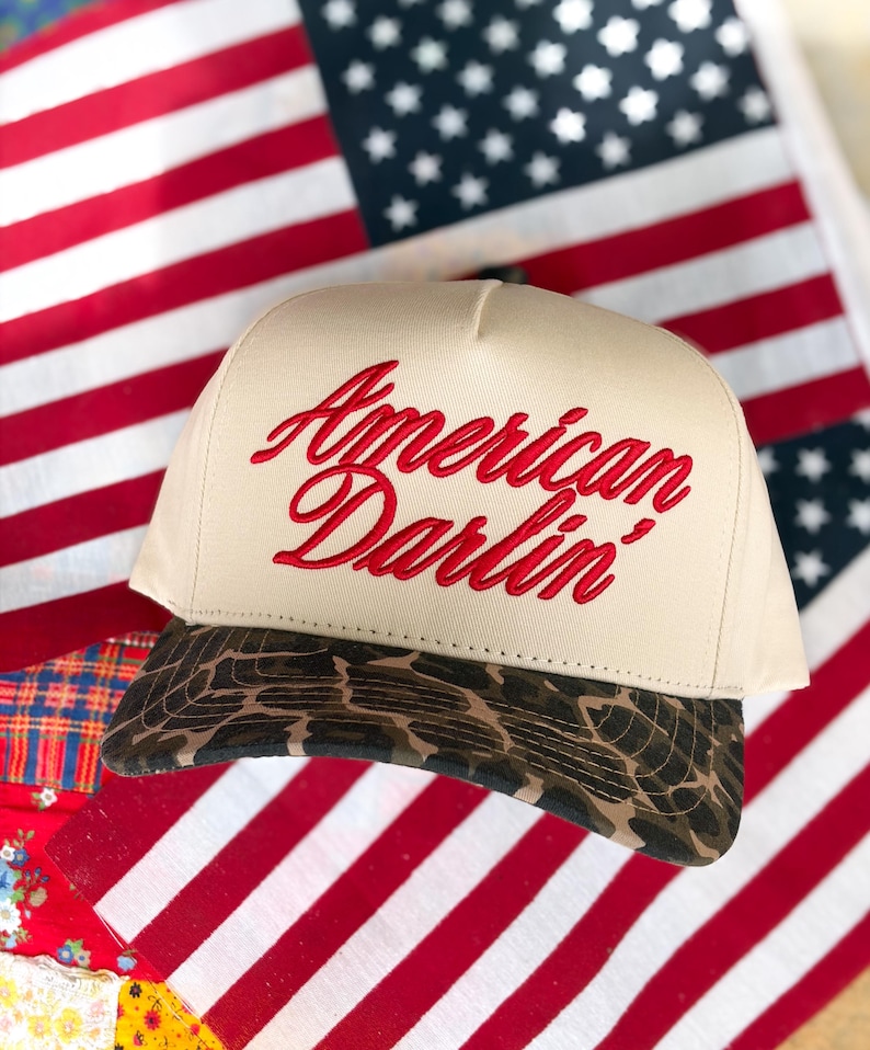 Puede incluir: Una gorra beige tipo trucker con el texto American Darlin' bordado en rojo en el panel frontal. La gorra tiene una corona estructurada y una visera curva con un patr&oacute;n de estampado animal marr&oacute;n y negro, sobre una bandera roja, blanca y azul.