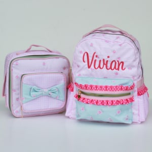 May include: Pink and blue floral print backpack with a matching lunch bag. The backpack has a pink ruffle trim and a zippered pocket with a gold zipper pull. The lunch bag has a gingham pattern and a bow. The backpack is personalized with the name "Vivian" in red embroidery.