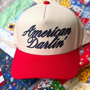 Puede incluir: Una gorra de b&eacute;isbol beige con visera roja brillante y un peque&ntilde;o bot&oacute;n rojo en la parte superior. El panel frontal presenta el texto American Darlin' bordado en cursiva azul marino oscuro. La gorra tiene una corona estructurada y visera.