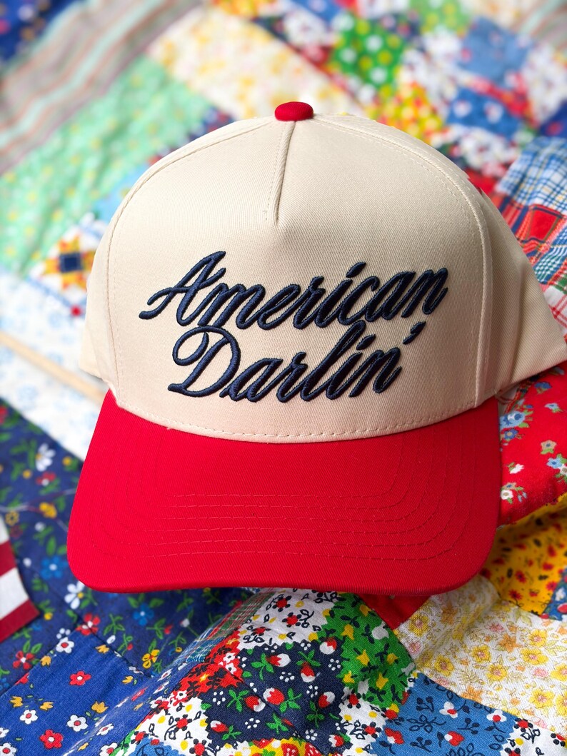 Puede incluir: Una gorra de b&eacute;isbol beige con visera roja brillante y un peque&ntilde;o bot&oacute;n rojo en la parte superior. El panel frontal presenta el texto American Darlin' bordado en cursiva azul marino oscuro. La gorra tiene una corona estructurada y visera.