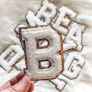 May include: White, fluffy fabric letter patches with gold glitter borders. The letter "B" is held in a hand, with other letters visible in the background. These patches are likely for personalization on clothing or accessories.
