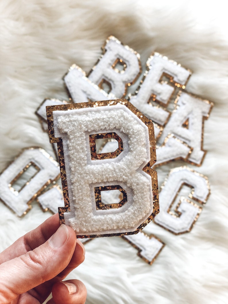 May include: White, fluffy fabric letter patches with gold glitter borders. The letter "B" is held in a hand, with other letters visible in the background. These patches are likely for personalization on clothing or accessories.