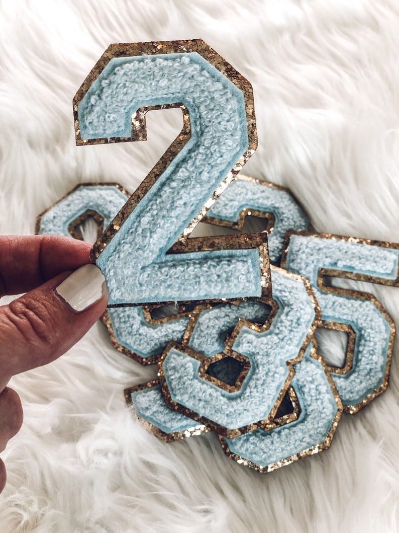 May include: Light blue chenille number patches with gold glitter borders. The number 2 is held up, with other numbers in the background. The patches have a soft, textured appearance, perfect for adding to clothing or accessories.