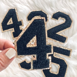May include: Black felt number patches with gold glitter edging. The numbers 4, 45, 2, and 5 are visible. The patches are displayed on a white, fluffy surface. These patches are ideal for adding to clothing or accessories.