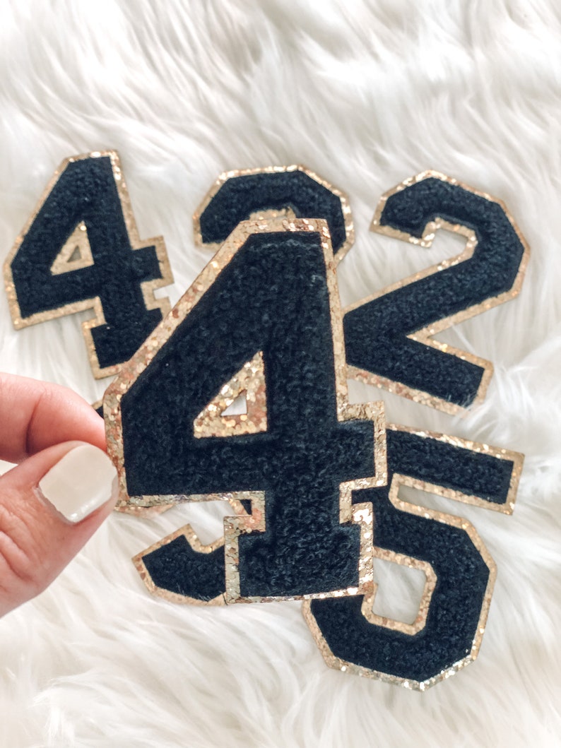 May include: Black felt number patches with gold glitter edging. The numbers 4, 45, 2, and 5 are visible. The patches are displayed on a white, fluffy surface. These patches are ideal for adding to clothing or accessories.