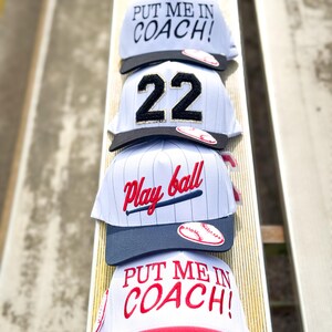 May include: Four baseball caps stacked on a wooden surface. The caps are white, gray, and red, with various designs including the text "PUT ME IN COACH!" and "Play ball". One cap displays the number "22".