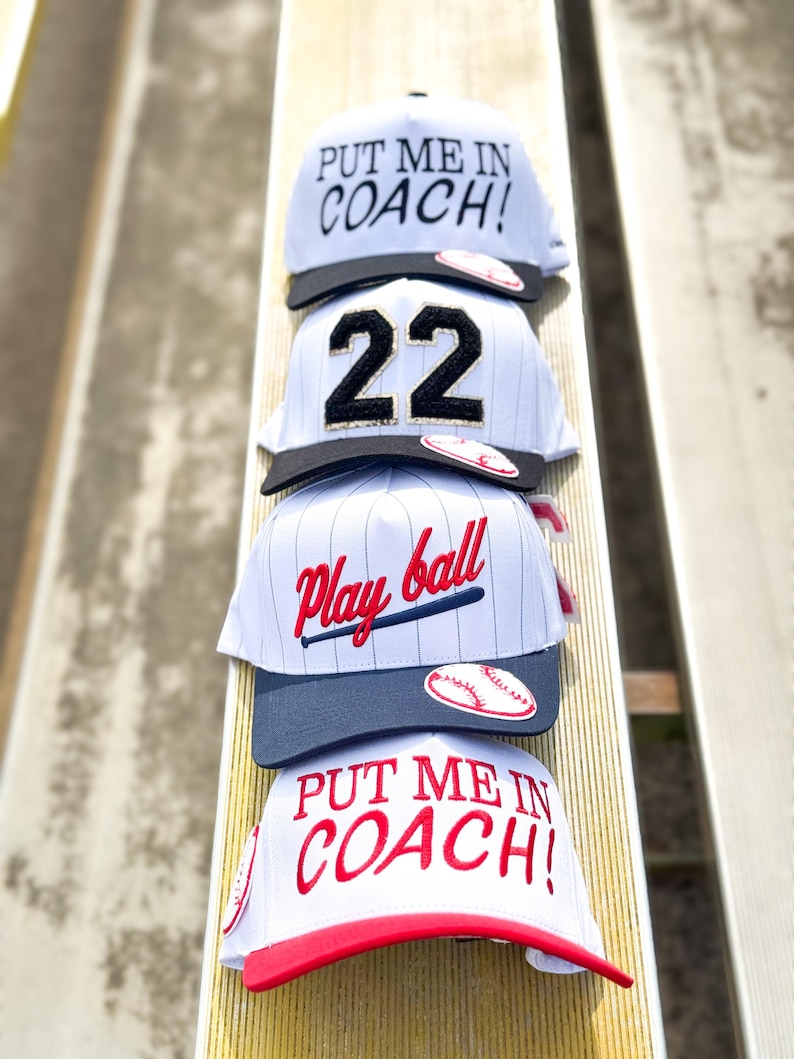May include: Four baseball caps stacked on a wooden surface. The caps are white, gray, and red, with various designs including the text "PUT ME IN COACH!" and "Play ball". One cap displays the number "22".