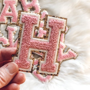 May include: A pink, gold, and white letter "H" patch with a textured, chenille-like appearance. The patch has a gold glitter border and a white inner border. Other letter patches are visible in the background.