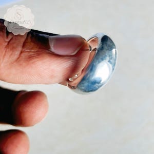 May include: A polished silver band ring with a smooth, rounded surface and a reflective finish. The metal has a bright, metallic lustre that catches the light. The ring is held in place, showcasing its simple, solid construction and curved profile.