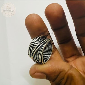 Puede incluir: Un anillo ancho de plata con un diseño texturizado de varias capas, compuesto por numerosos hilos finos superpuestos que envuelven la banda. El metal tiene un acabado pulido con áreas hundidas oscurecidas para resaltar el patrón intrincado.