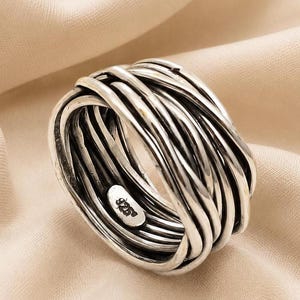 May include: A wide silver ring featuring a multi-strand, intertwined wire design that creates a textured, layered appearance. The interior of the band is clearly stamped with the hallmark 925, indicating the sterling silver composition of the piece.