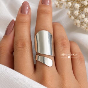 Puede incluir: Un anillo de plata pulida ancho con un diseño envolvente abierto, suave y curvado que cubre una parte importante del dedo. El anillo tiene una superficie metálica reflectante e incluye el texto silvoraa925 con un pequeño icono de corazón.