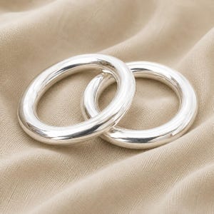 Puede incluir: Dos anillos plateados pulidos con un perfil circular grueso, liso y redondeado. Los anillos están entrelazados, mostrando una superficie metálica reflectante que capta la luz, creando una estética limpia y sencilla para estas piezas de joyería.