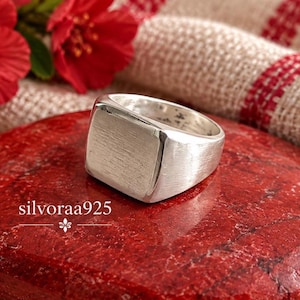 Peut inclure: Une bague chevalière en argent avec une finition brossée et mate sur la face carrée et un anneau poli. La bague présente une surface supérieure plate et rectangulaire avec le texte silvoraa925 imprimé en blanc sur le côté inférieur gauche.