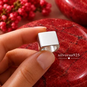 May include: A polished silver ring featuring a flat, square-shaped top surface. The ring is held against a background of red textured stone and small red berries. The text silvoraa925 is printed in white on the lower right side of the image.