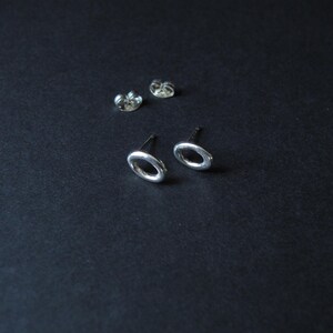 May include: A pair of silver stud earrings featuring a simple oval or circular hoop design. The earrings include two matching metal earring backs, all displayed with a polished metallic finish that reflects light against the dark surface.