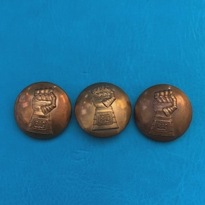 May include: Three circular bronze-toned buttons, each featuring a raised embossed design of a gauntlet or armoured fist. The base of each gauntlet displays the letters RAC in a rectangular frame. The buttons have a smooth, metallic finish.