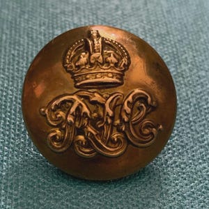 May include: A circular, metallic, bronze-toned button featuring a raised, embossed design. The top portion displays a detailed crown, while the lower portion features the intertwined, ornate, and stylized letters R and C in a decorative script.