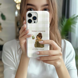 Puede incluir: Una funda para teléfono con un patrón de cuadros vichy en verde claro y blanco. El centro presenta la ilustración de una persona con un moño sosteniendo una taza, una vela pequeña y el texto "TODAY I CHOOSE TO SLOW DOWN" en letras marrones.
