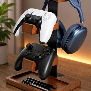 May include: A gaming accessory stand featuring a black metal vertical post with two wooden controller mounts and a top wooden headphone hanger. The base is a rectangular wooden tray holding three pens. A white controller, a black controller, and blue headphones are displayed.