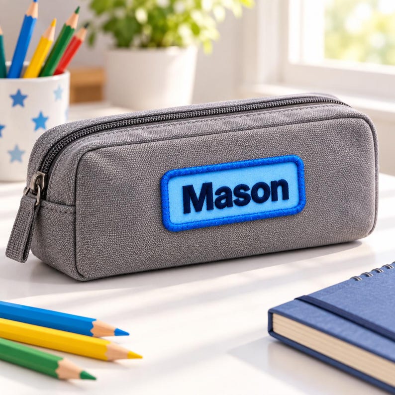 May include: A rectangular gray canvas pencil case with a zipper closure. Centered on the front is a rectangular light blue patch with a dark blue border, featuring the name Mason in bold, dark blue, sans-serif capital and lowercase letters.