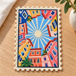 May include: A rectangular embroidered patch shaped like a postage stamp featuring a vibrant scene of colorful buildings surrounding a central sun with radiating light beams. The design includes yellow, pink, orange, and blue houses with green foliage.