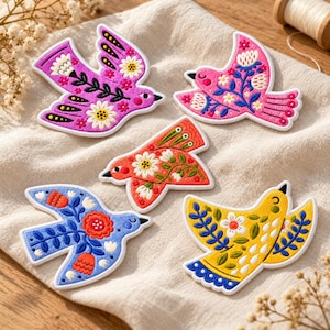 May include: Five embroidered bird-shaped patches featuring vibrant colors including purple, pink, orange, blue, and yellow. Each patch displays intricate floral patterns, white outlines, and black beaks, arranged on a textured off-white fabric surface.