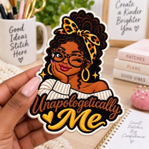 May include: An embroidered patch featuring a person with dark skin, curly hair, and a leopard print headwrap. The person wears glasses and gold hoop earrings, with the text "Unapologetically Me" in gold and brown script at the bottom of the patch.