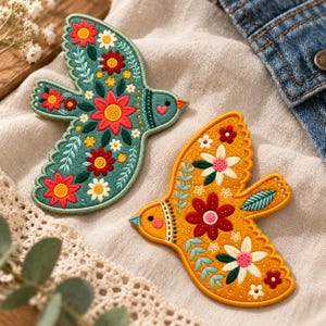 May include: Two embroidered bird-shaped patches. One patch is teal with red, yellow, and white floral patterns. The other patch is mustard yellow with red, white, and teal floral and leaf designs. Both birds feature small pink hearts on their cheeks.