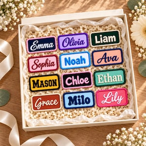 May include: A collection of twelve rectangular embroidered name patches in a gift box. The patches feature various colors and fonts with the names Emma, Olivia, Liam, Sophia, Noah, Ava, Mason, Chloe, Ethan, Grace, Milo, and Lily stitched on them.