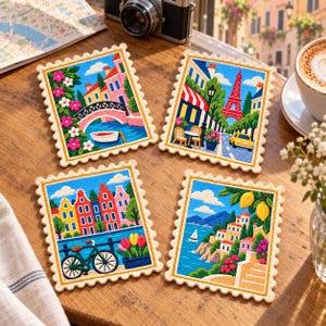 May include: Four rectangular coasters with scalloped edges, each featuring a colorful embroidered scene: a canal bridge with flowers, a Parisian street with the Eiffel Tower, a row of Dutch houses with a bicycle, and a coastal Mediterranean village.
