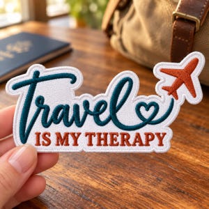 May include: An embroidered patch with a white background and a dark teal script that reads Travel, followed by a small heart and an orange airplane icon. Below the script, the text IS MY THERAPY is embroidered in orange block letters.