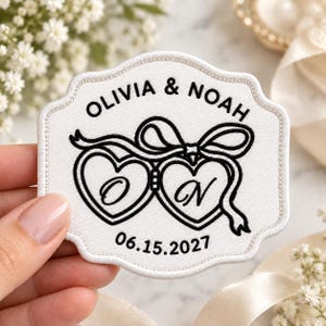 May include: A white embroidered patch with a scalloped edge featuring black stitching. The design includes the text OLIVIA & NOAH, two connected heart outlines with the letters O and N inside, a decorative ribbon bow, and the date 06.15.2027.