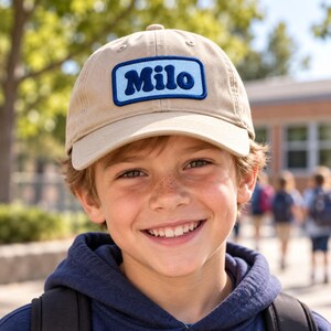 K&ouml;nnte beinhalten: Eine beige Baseballkappe mit einem rechteckigen blauen Aufn&auml;her auf der Vorderseite. Der Aufn&auml;her hat einen hellblauen Rand und zeigt das Wort Milo in einer fetten, dunkelblauen, abgerundeten Schrift. Die Kappe hat einen gebogenen Schirm.