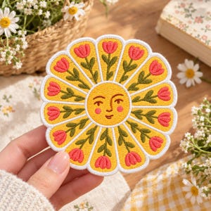 May include: A circular embroidered patch featuring a central yellow sun face with a serene expression. The sun is surrounded by twelve yellow petals, each containing a green stem with a single pink tulip flower, all outlined in white thread.