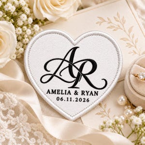May include: A white heart-shaped embroidered patch featuring black stitched lettering with the monogram AR, the names AMELIA & RYAN, and the date 06.11.2026. The patch has a white stitched border and a textured fabric surface.