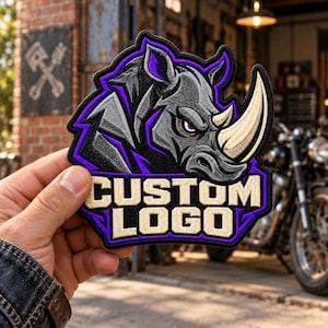 May include: A fabric patch featuring an embroidered graphic of a gray rhinoceros head with a large horn, accented by purple outlines. Below the rhinoceros, the words CUSTOM LOGO are embroidered in beige block letters on a black background.