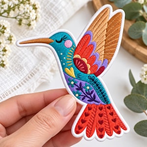 May include: An embroidered hummingbird patch featuring a vibrant color palette of teal, orange, red, purple, and yellow. The design includes intricate stitching details such as small flowers, heart shapes, and a white border outline.