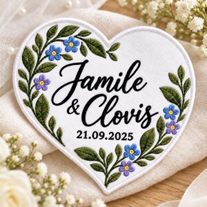 May include: A heart-shaped white fabric patch featuring black embroidered text that reads Jamile & Clovis and 21.09.2025. The patch is decorated with an embroidered border of green leaves and small blue and purple flowers.