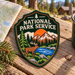May include: A shield-shaped embroidered patch featuring the text NATIONAL PARK SERVICE in white. The design depicts a brown elk standing near a blue lake, green pine trees, snow-capped mountains, and an orange sun against a dark green background.
