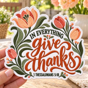 May include: A white embroidered patch featuring the text IN EVERYTHING give thanks 1 THESSALONIANS 5:18 in orange and black lettering. The design is surrounded by orange and peach tulips with dark green leaves and small orange berries.