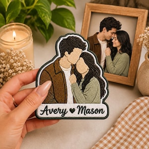May include: A custom embroidered patch featuring a stylized illustration of two people embracing. The patch displays a person in a brown jacket and a person in a green sweater, with the text Avery ♥ Mason embroidered in black on a white base.