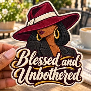 May include: An embroidered patch featuring a person wearing a wide-brimmed burgundy hat and a black blazer, accented with gold hoop earrings. The text "Blessed and Unbothered" is stitched in cream and gold lettering at the bottom of the patch.