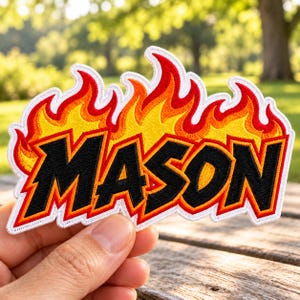 May include: An embroidered patch featuring the name MASON in bold black letters with a thick red and orange outline. The text is set against a vibrant, stylized flame design in shades of yellow, orange, and red, finished with a white border.