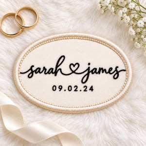 May include: An oval-shaped cream-colored fabric patch with a gold embroidered border. The patch features black embroidered text that reads "sarah & james" with a heart symbol in the center, followed by the date "09.02.24" in a simple black font.