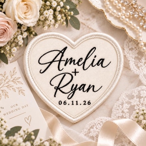 May include: A heart-shaped fabric patch with a cream-colored base and a stitched border. The patch features black embroidered text that reads Amelia + Ryan and the date 06.11.26 in a clean, sans-serif font below the names.